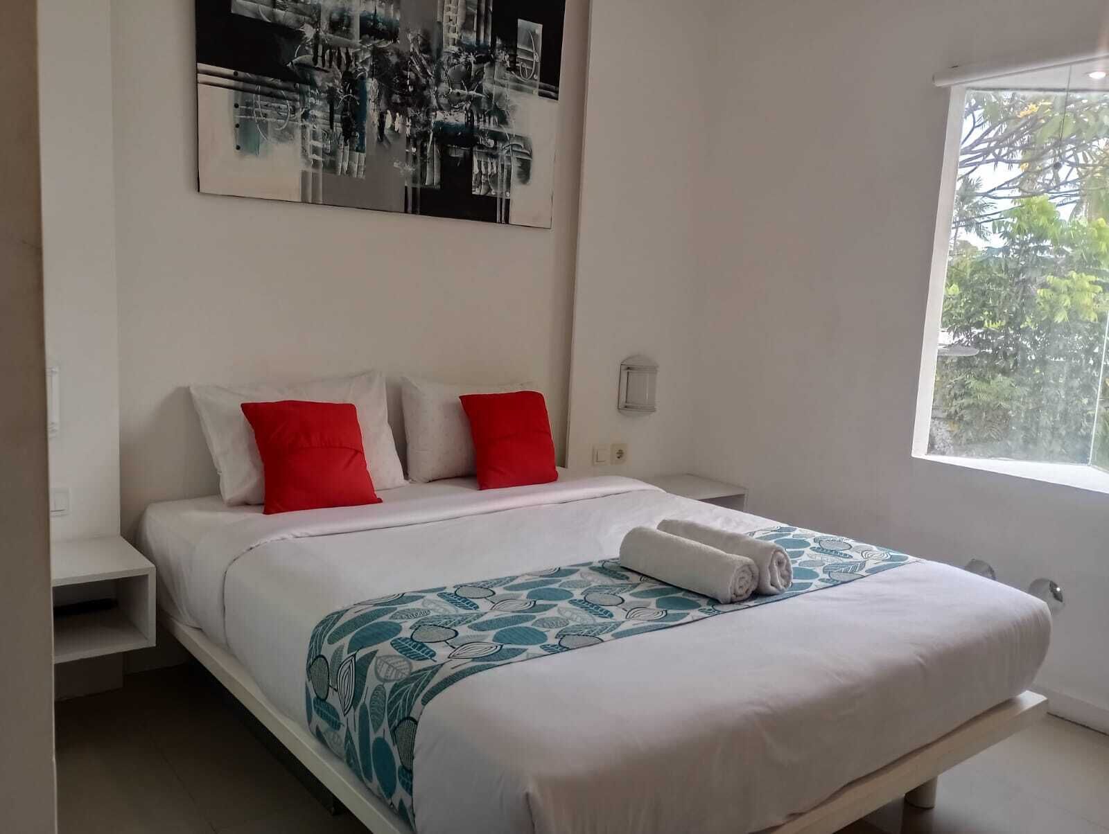 Quarto com cama, mesa de cabeceira, luminária na parede e janela com vista para árvores.