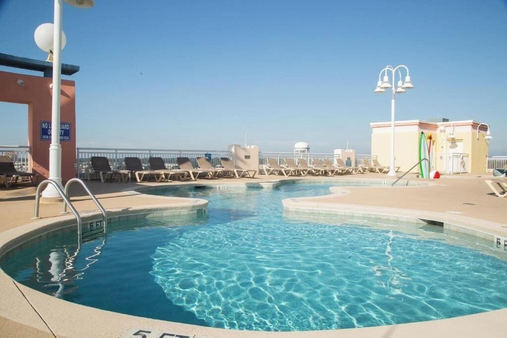 Ein Poolbereich mit Liegestühlen, einem Rettungsschwimmerstand und blauem Himmel.