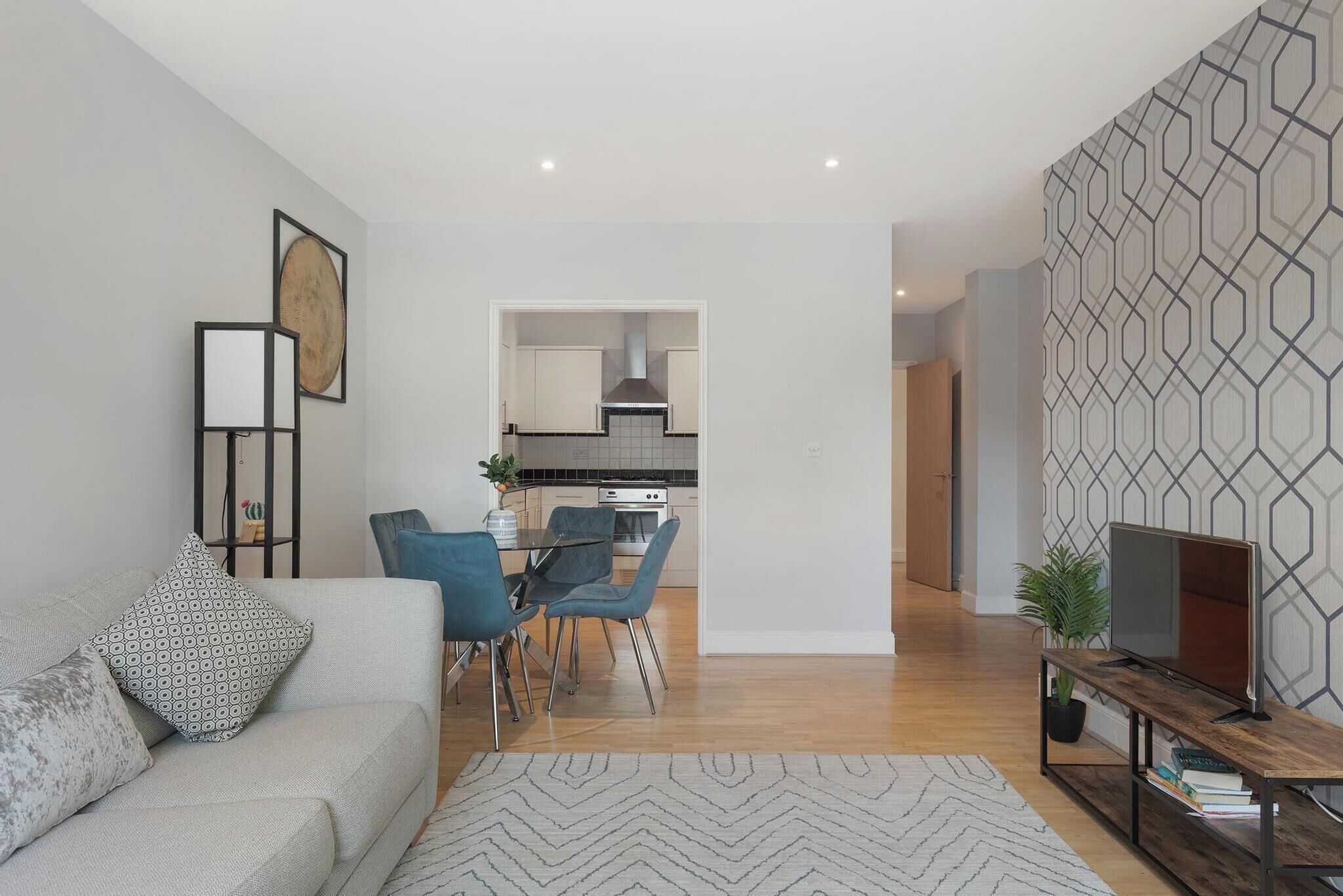 Una sala moderna con un sofá, cojines estampados, una zona de comedor con sillas azules, una cocina al fondo y un televisor sobre un mueble de madera.