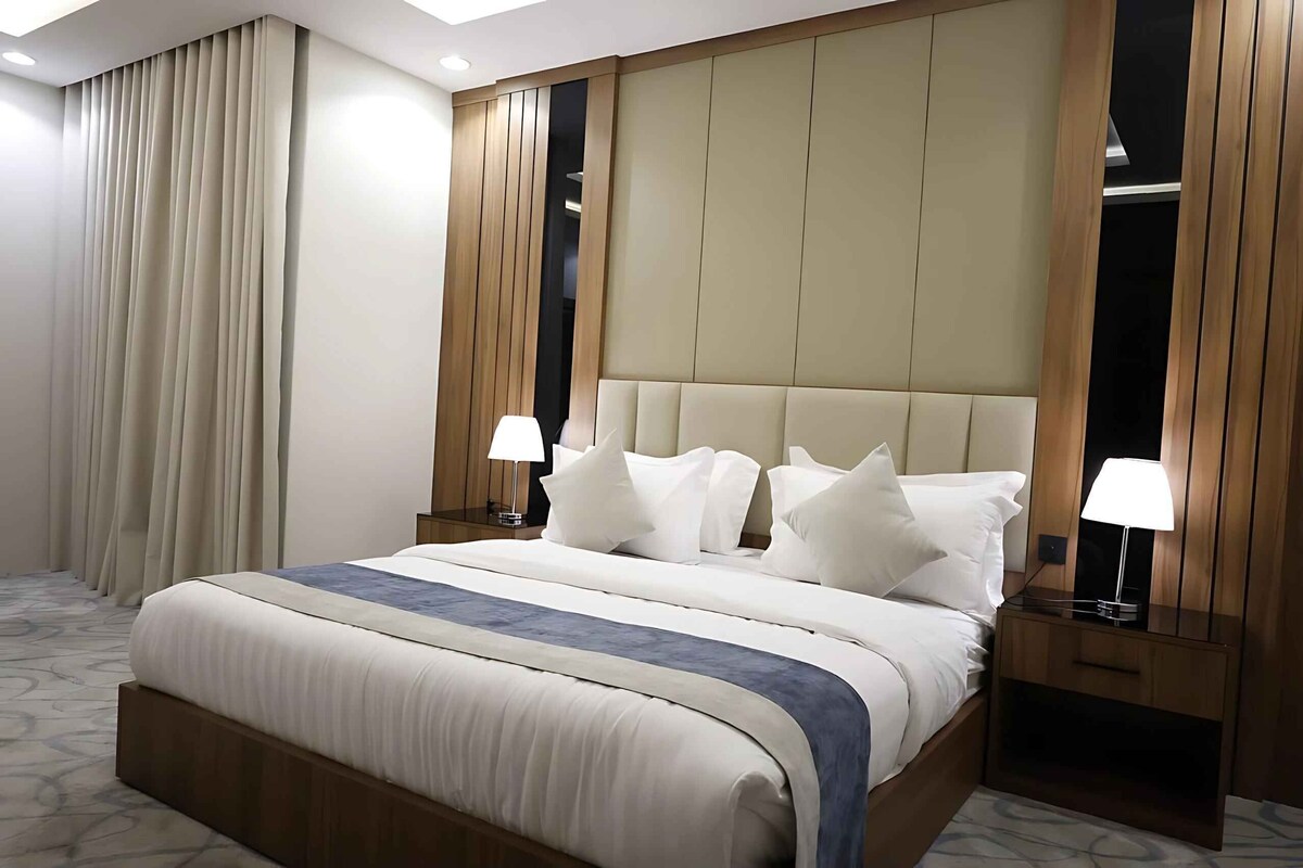 A hotel room with a large bed, two bedside tables with lamps, a headboard, and a view of the outside through a window with curtains.