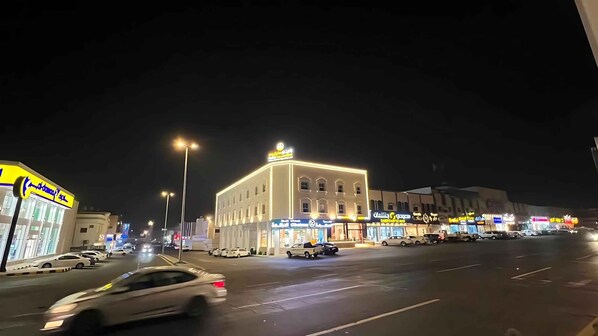 A nighttime street scene with a building featuring multiple storefronts, a parking area, and several cars.