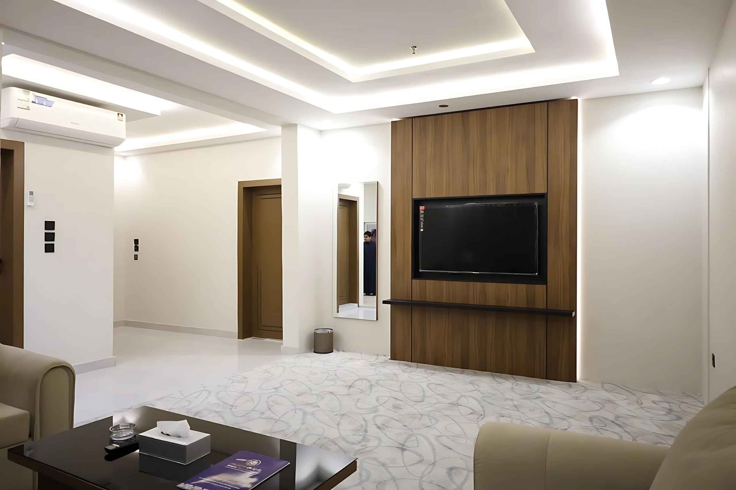 A modern hotel room with a flat-screen TV mounted on a wooden panel, a marble-patterned floor, and a sofa.