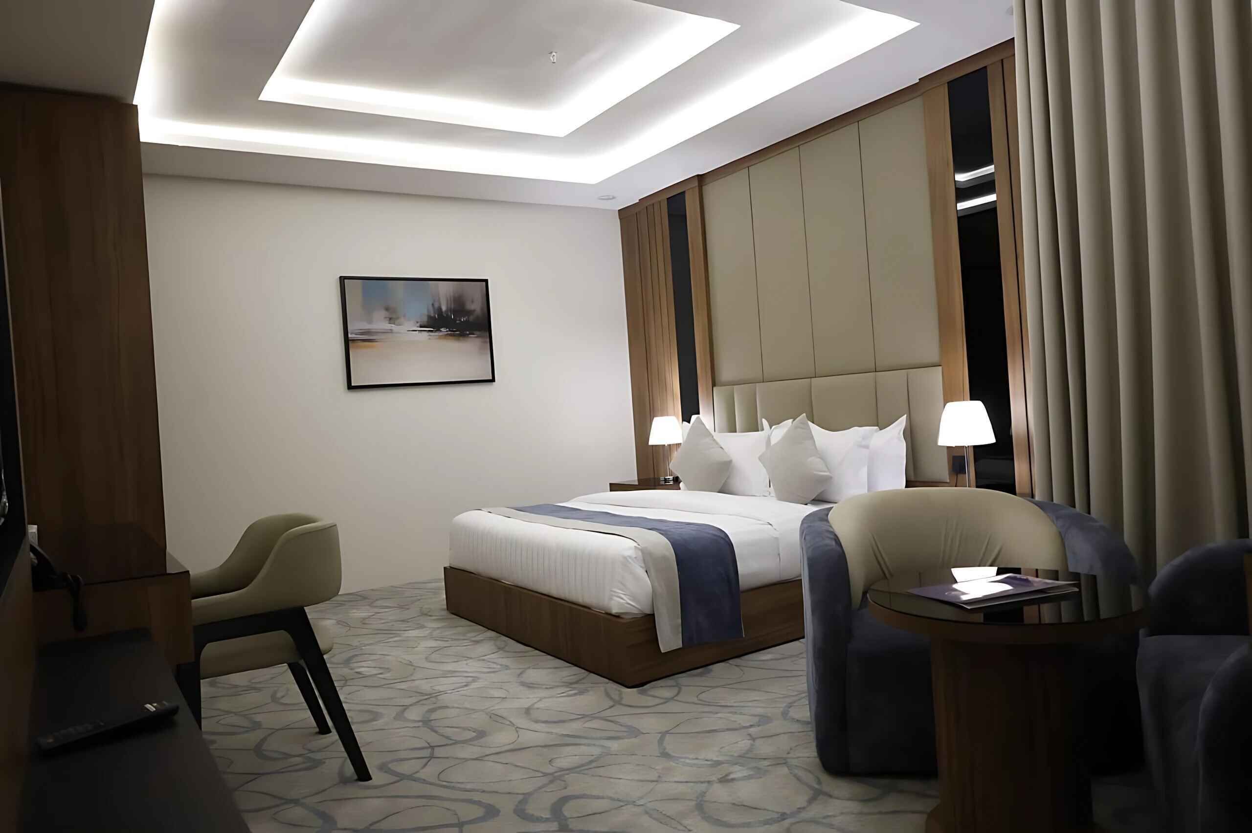 Una habitación de hotel con una cama, dos lámparas de noche, una silla, una mesita y un cuadro en la pared.