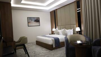 Una habitación de hotel con una cama, dos lámparas de noche, una silla, una mesita y un cuadro en la pared.