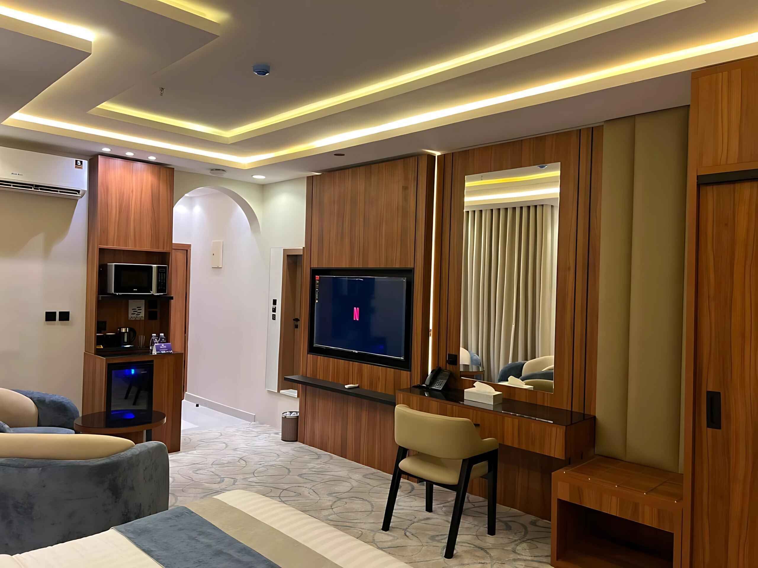 Una habitación de hotel moderna con televisor de pantalla plana, paneles de madera, una zona de estar con sofá y sillón, y un escritorio pequeño con una silla.