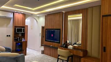 Una habitación de hotel moderna con televisor de pantalla plana, paneles de madera, una zona de estar con sofá y sillón, y un escritorio pequeño con una silla.