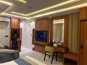 A modern hotel room with a flat-screen TV, wooden paneling, a seating area with a sofa and armchair, and a small desk with a chair. - RADIX MILLENNIUM (Al-Rawna District)