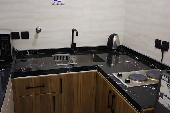 A compact kitchen with a black countertop, a sink, a kettle, and a stove. - RADIX MILLENNIUM (Al-Rawna District)