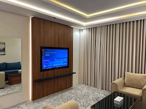 Una sala de estar moderna con una televisión de pantalla plana montada en un panel de madera, un sofá, una mesa de centro y un ventanal con cortinas.