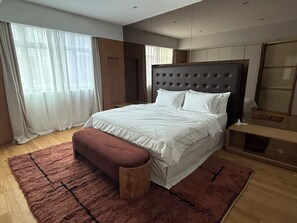 A modern bedroom with a large bed, a wooden bench, and a view of the city through large windows.