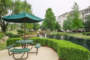 A picnic table with a green umbrella, surrounded by well-manicured hedges and a pond with a bridge. - Spacious 2BR Suite Near Lake & Attractions (Branson)