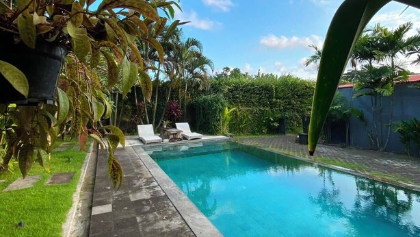 A rectangular swimming pool with a clear blue surface, surrounded by a paved deck and a grassy area. There are lounge chairs and a small table near the pool's edge. - Pondok Cintya Kerobokan Canggu (Badung)