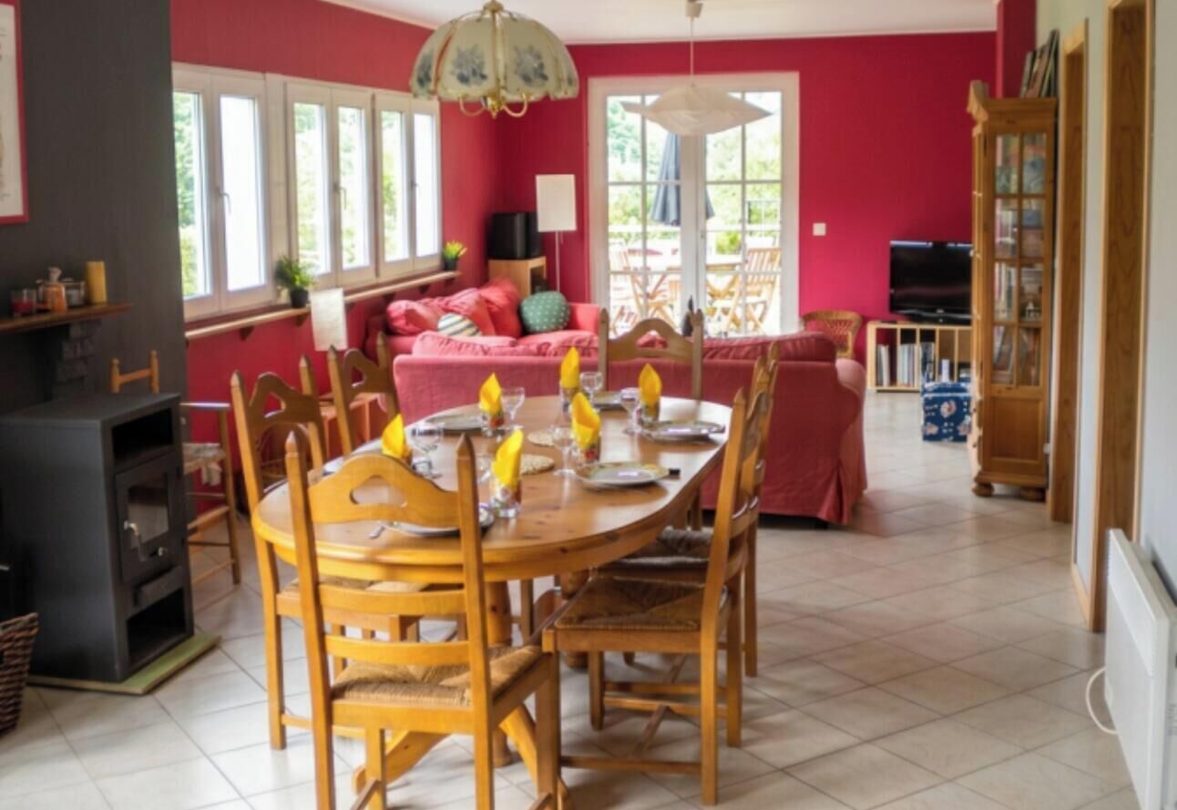 Un coin repas avec une table ronde dressée pour quatre personnes, entourée de chaises en bois. On aperçoit un canapé et un téléviseur au fond.