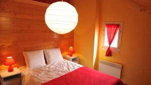 Ein Schlafzimmer mit hölzernem Kopfteil, einem Bett mit weißer Bettwäsche, einer roten Tagesdecke, zwei Nachttischlampen und einem Fenster mit rotem Vorhang.