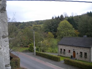 A rural road leading to a stone building with a sloped roof, surrounded by dense green trees and a hill in the background. - Le Chênelierre - Everything for the Kids - Dogs Welcome (Free) (Manhay)