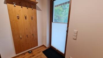 Een kamer met een houten garderobe, een zwarte mat bij de ingang, en een raam met zonwering.