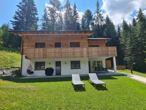 A two-story wooden house with a balcony, a covered patio, and a grassy lawn with two lounge chairs. - Chalet Rehblick (Bad Kleinkirchheim)