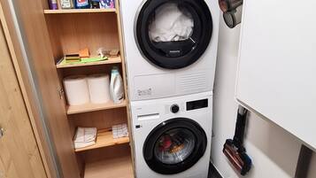 A compact laundry room with a front-loading washer and dryer, a shelf with cleaning supplies, and a laundry basket.