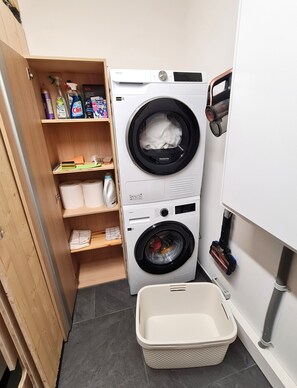 A compact laundry room with a front-loading washer and dryer, a shelf with cleaning supplies, and a laundry basket.