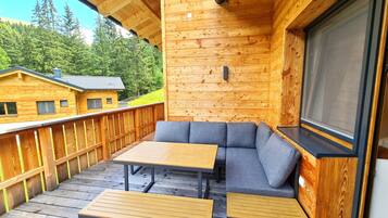Eine Holzterrasse mit einer Couch und Tischen, mit Blick auf ein bewaldetes Gebiet.