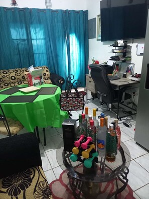 Ein Wohnzimmer mit einem grünen Tisch, einer Couch, einem Couchtisch mit Flaschen und Gewichten, einem Fernseher und einem Schreibtisch mit einem Computer.