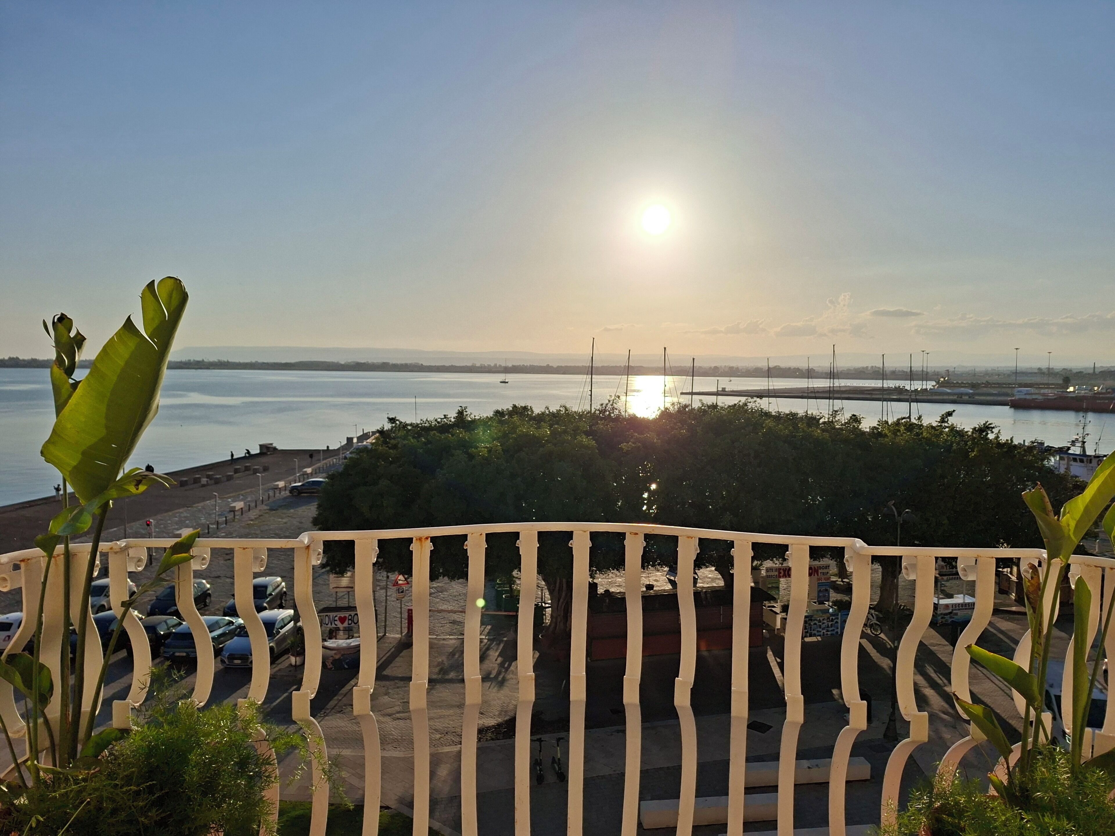 A view of a waterfront with a sun setting behind a bridge, a parking area, and a fence with greenery in the foreground.