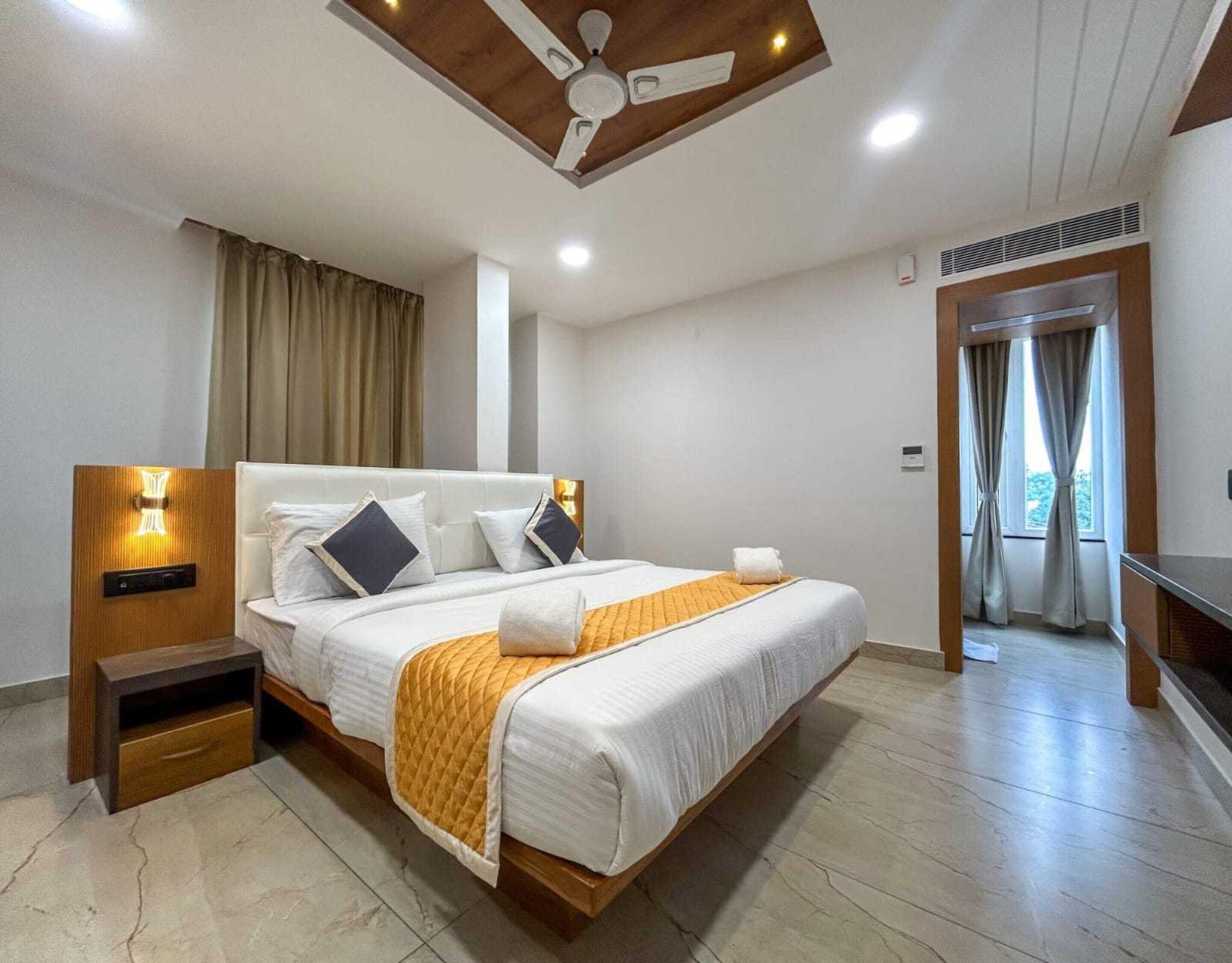 Ein Hotelzimmer mit einem Bett, Nachttischen, einem Deckenventilator, einem Fenster mit Vorhängen und einer Tür zu einem anderen Raum.