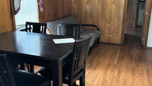 Una habitación con paneles de madera, una mesa de comedor y sillas, una ventana con cortinas y una puerta que da a otra habitación.