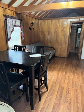 A wooden-paneled room with a dining table and chairs, a window with curtains, and a door leading to another room.
