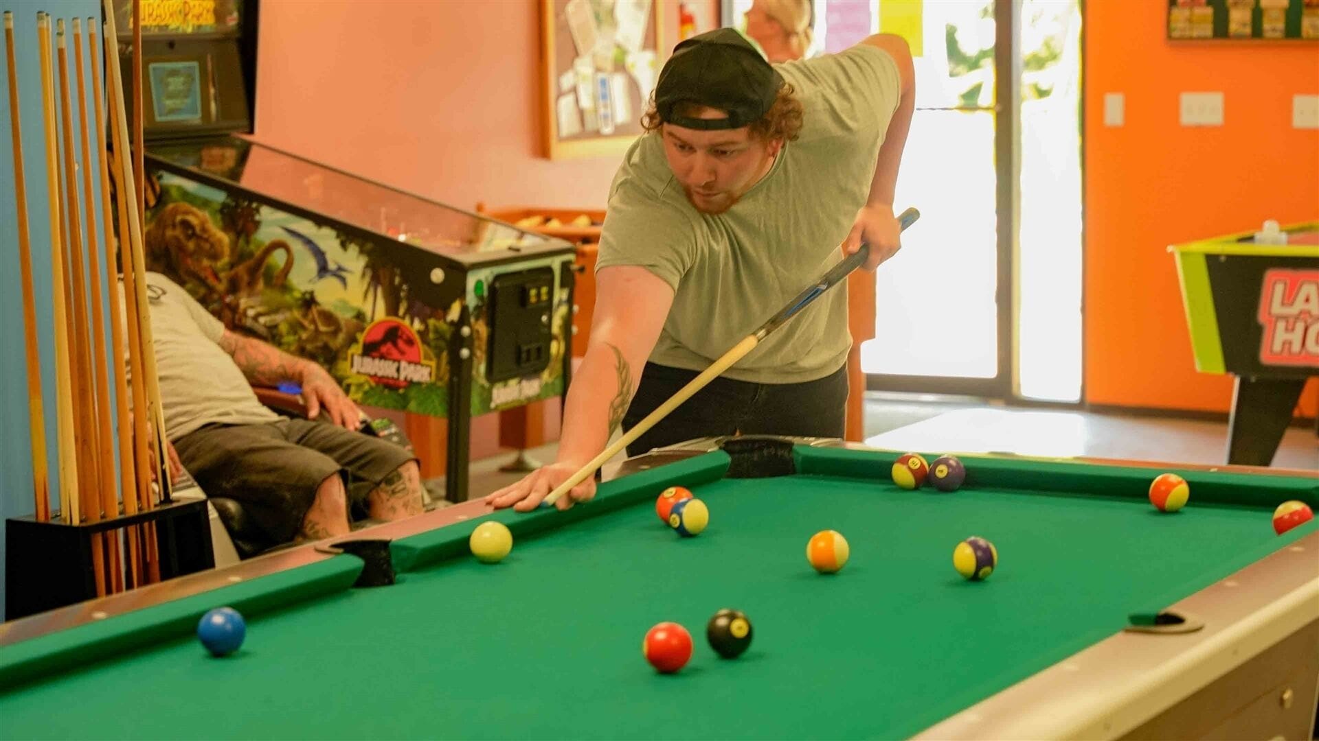 A pool table with balls and cues, a person playing, and a dinosaur-themed arcade machine in the background.