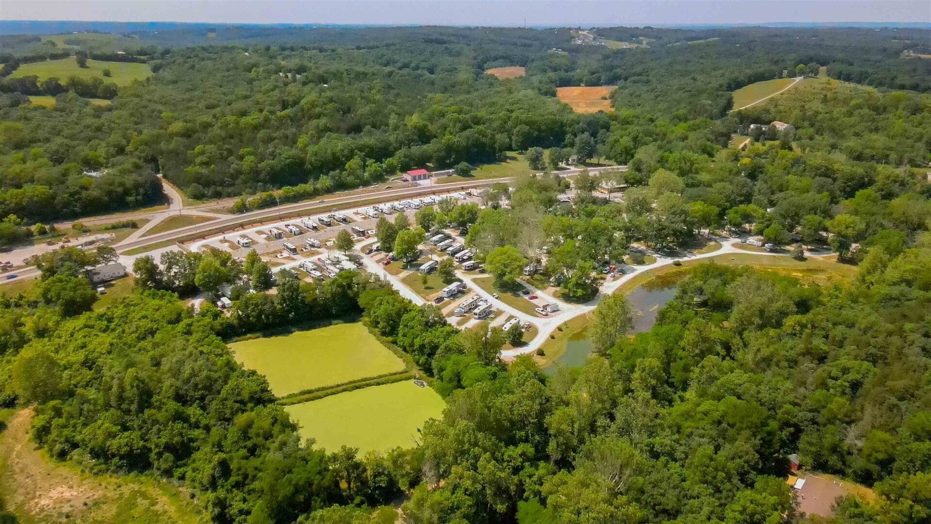 An aerial view of a campground with RV parking, a small lake, and surrounding wooded areas.