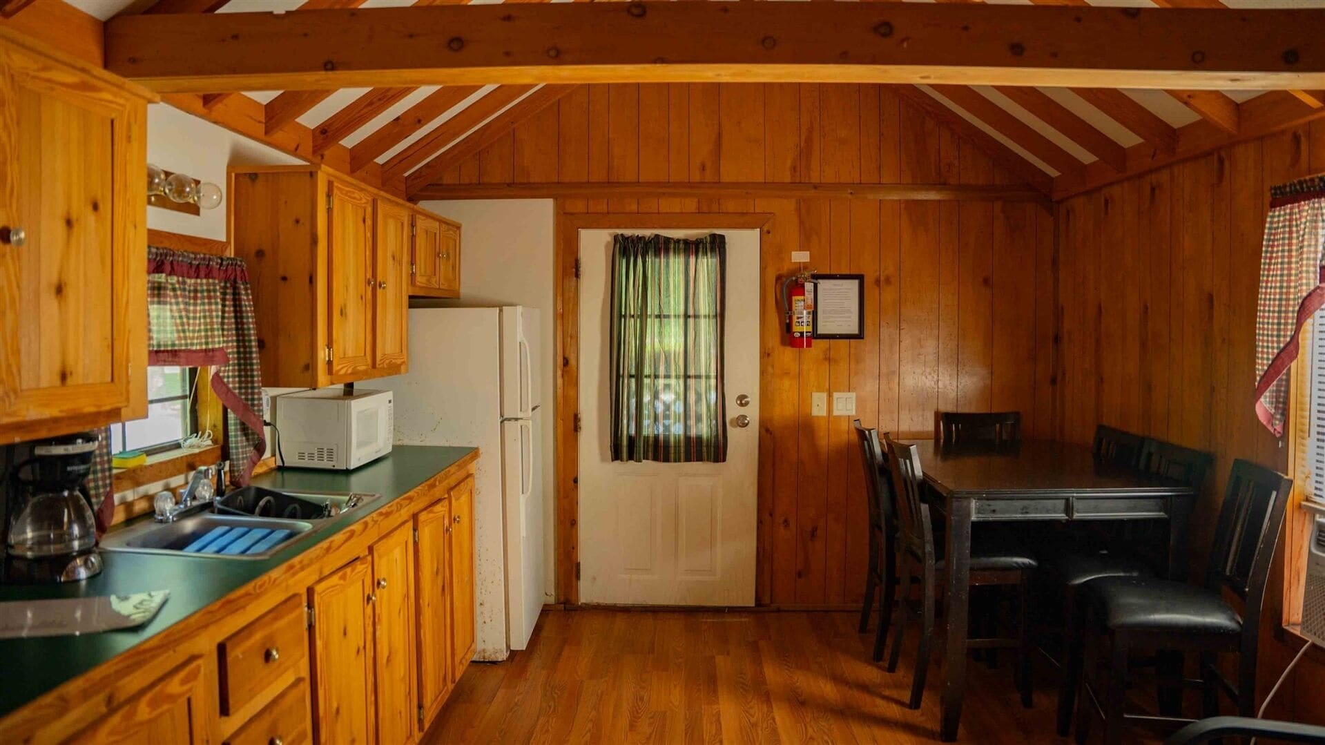 Cocina compacta con armarios de madera, microondas y refrigerador. Hay una mesa de comedor con sillas y una ventana con cortinas.