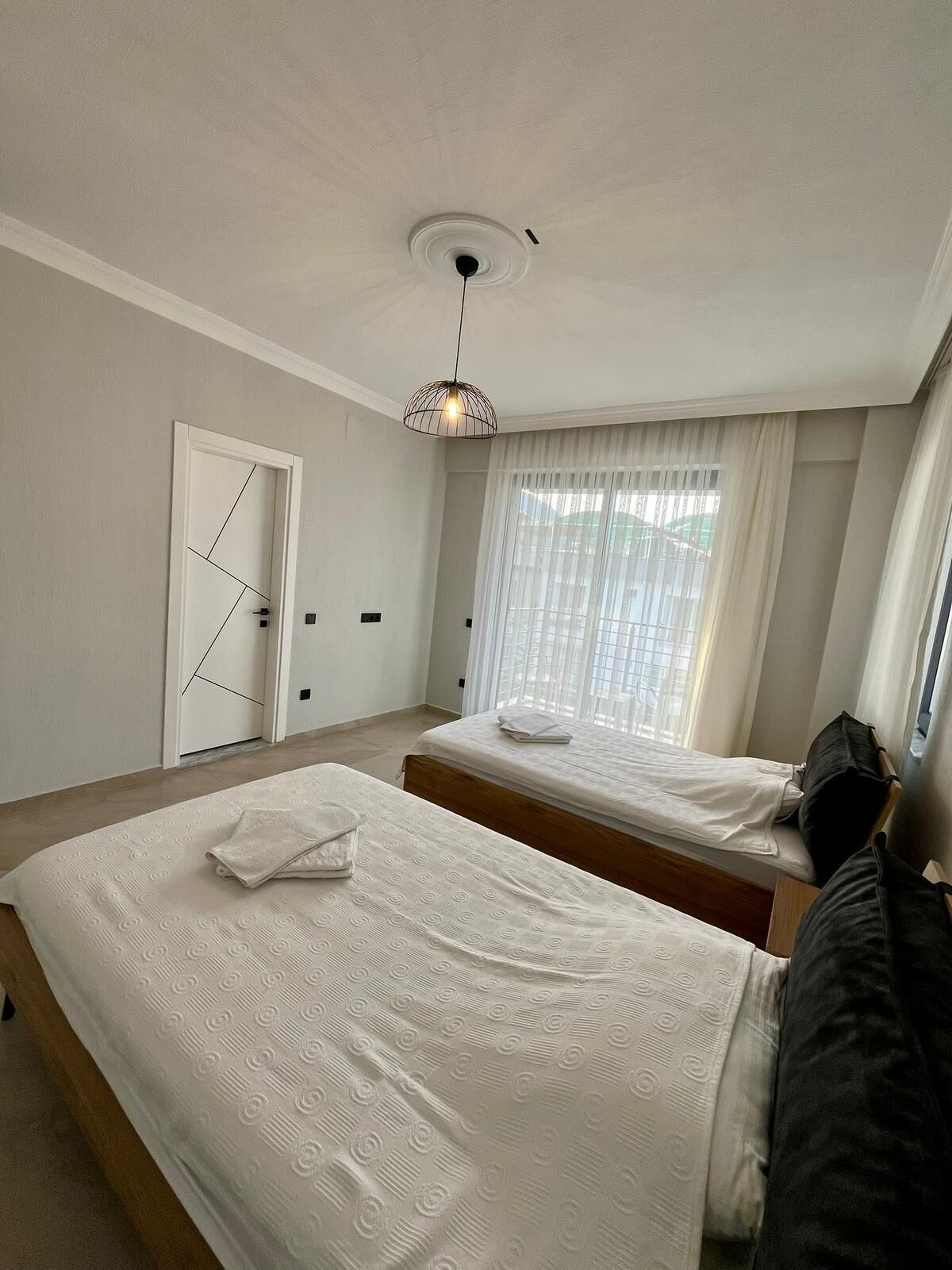 A hotel room with two beds, a ceiling light fixture, a window with curtains, and a door.
