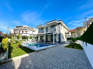 A modern two-story house with a swimming pool, surrounded by a well-maintained lawn and a gravel driveway.