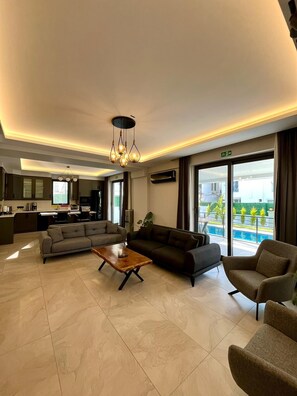 A modern hotel lobby with a seating area, a kitchen in the background, and large windows offering a view of the outdoors.