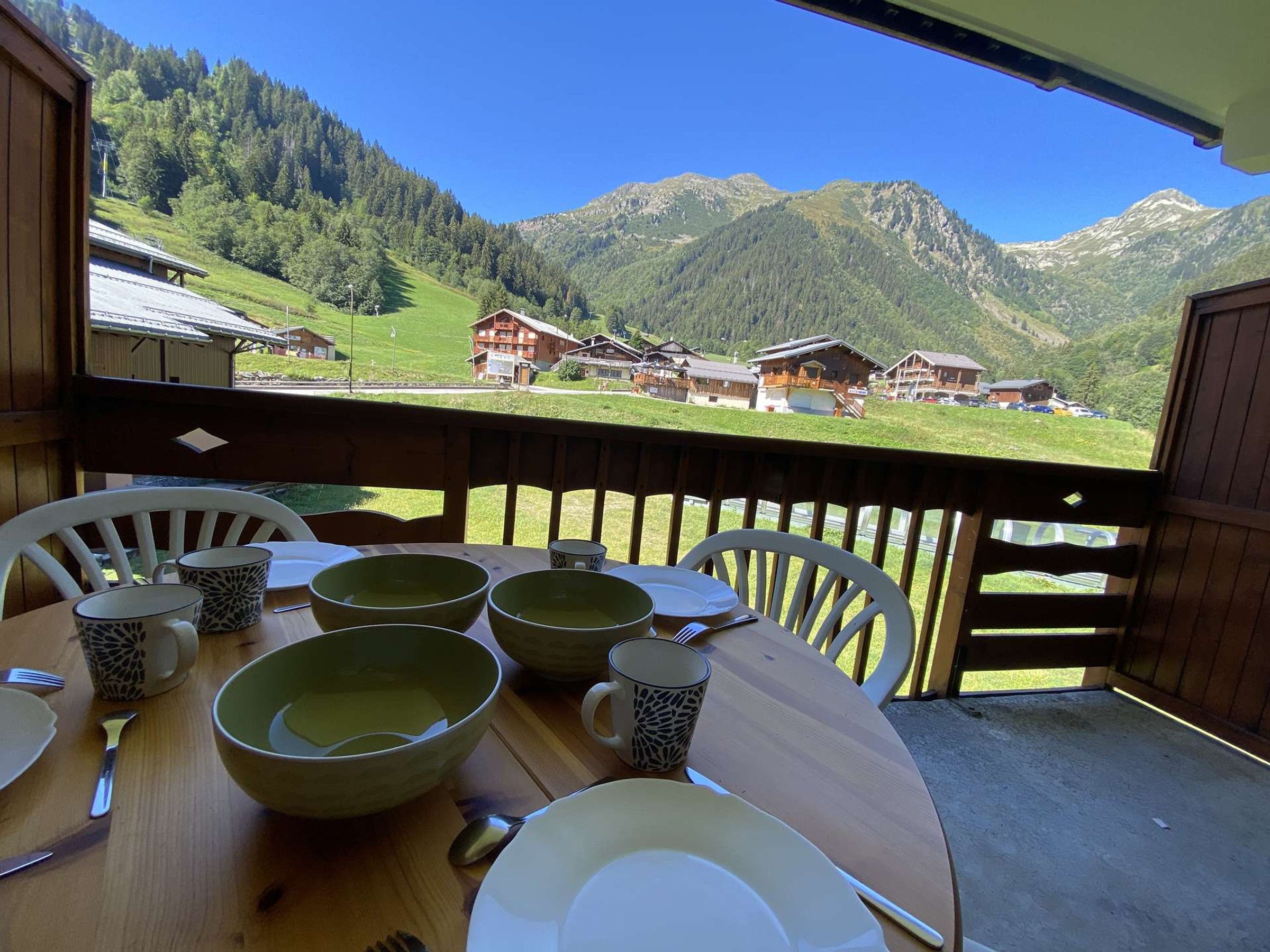 A dining table with chairs and tableware on a balcony overlooking a mountainous landscape with traditional buildings.