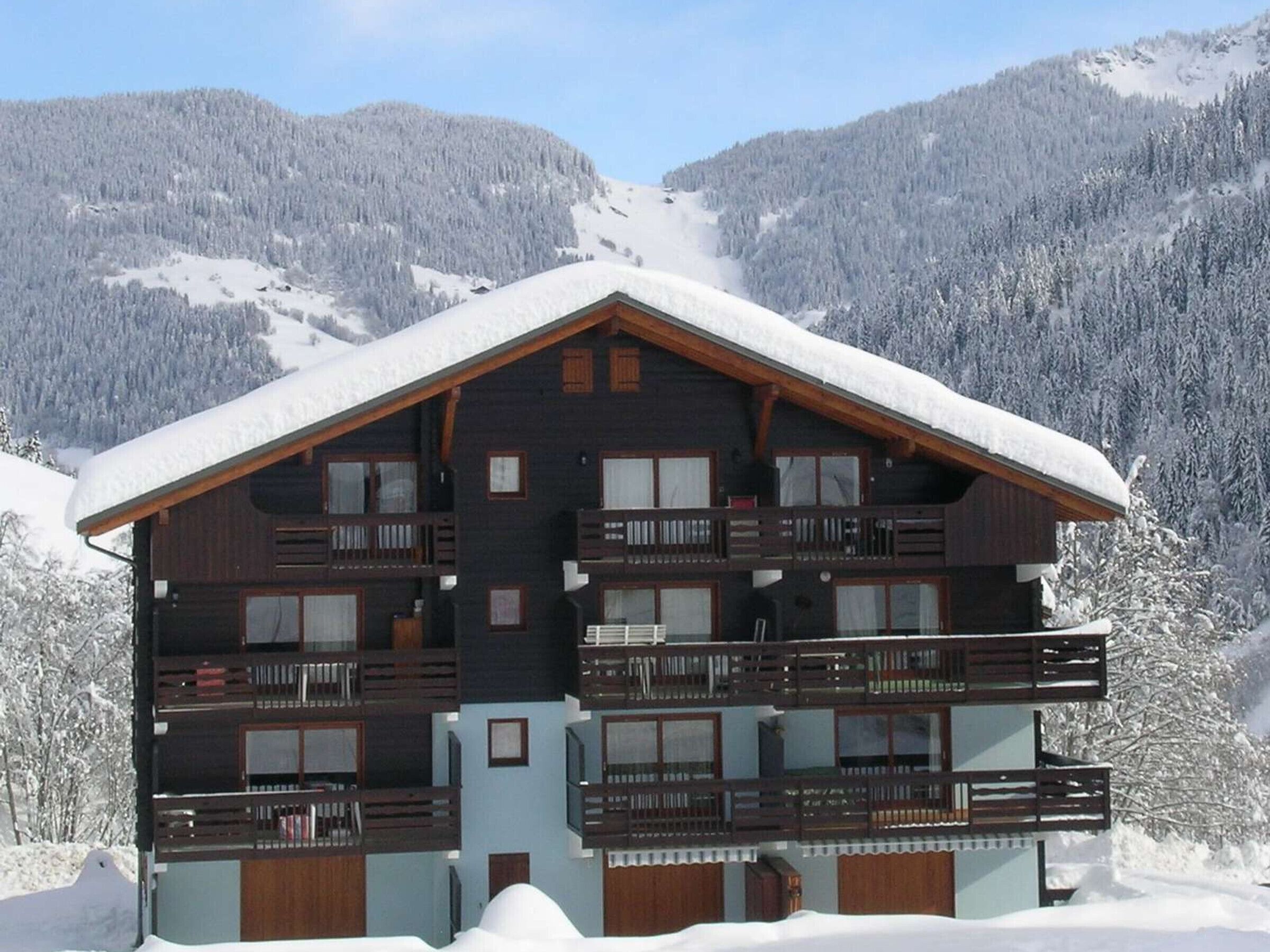 A multi-story wooden chalet with balconies, set against a snowy mountainous backdrop.