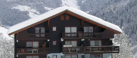 A multi-story wooden chalet with balconies, set against a snowy mountainous backdrop.