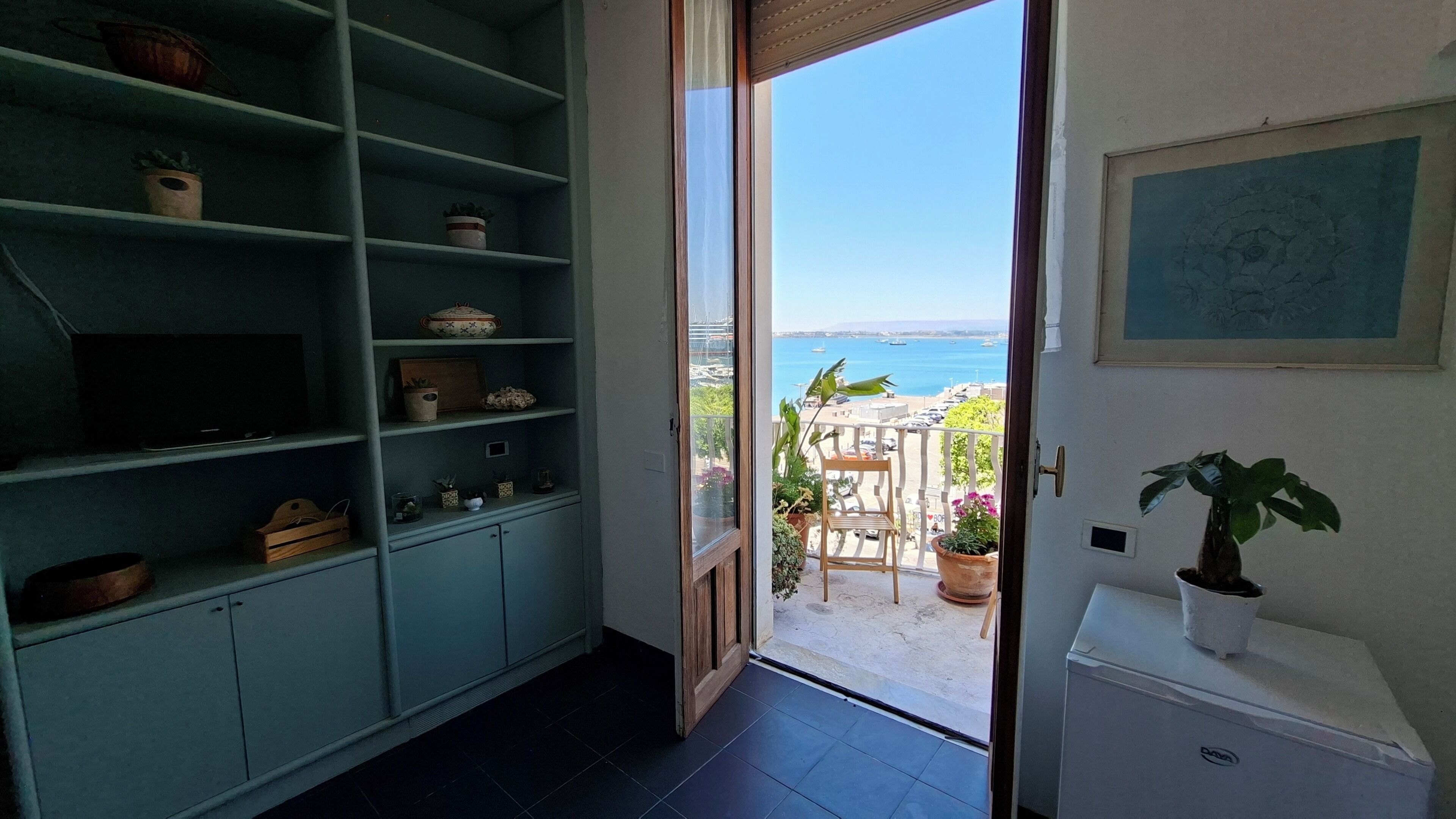 A room with a view of the sea, a balcony with a table and chairs, and a shelving unit with various items.