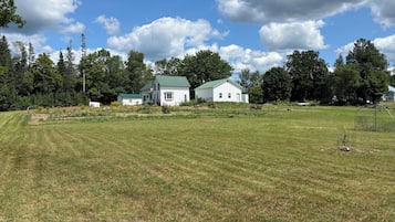 A grassy field with two white buildings, one with a green roof, surrounded by trees and a clear sky.