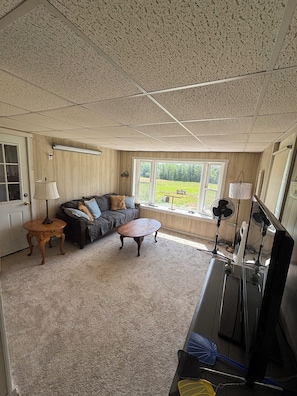 A living room with a sectional sofa, a coffee table, and a television. There is a window providing a view of a grassy area. - Unplug at Gentle Giant Acres — Historic Maine Farmhouse on 16 Acres (Kingman)