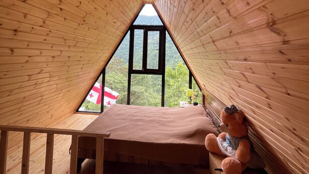 A triangular wooden room with a bed, a teddy bear, and a window overlooking greenery. - Machaxela Skyline Cottage – Scenic Mountain Getaway near Batumi. (Khelvachauri municipality)