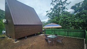 A wooden structure with a sloped roof, a fenced outdoor area with a table and chairs, and a colorful umbrella. - Machaxela Skyline Cottage – Scenic Mountain Getaway near Batumi. (Khelvachauri municipality)