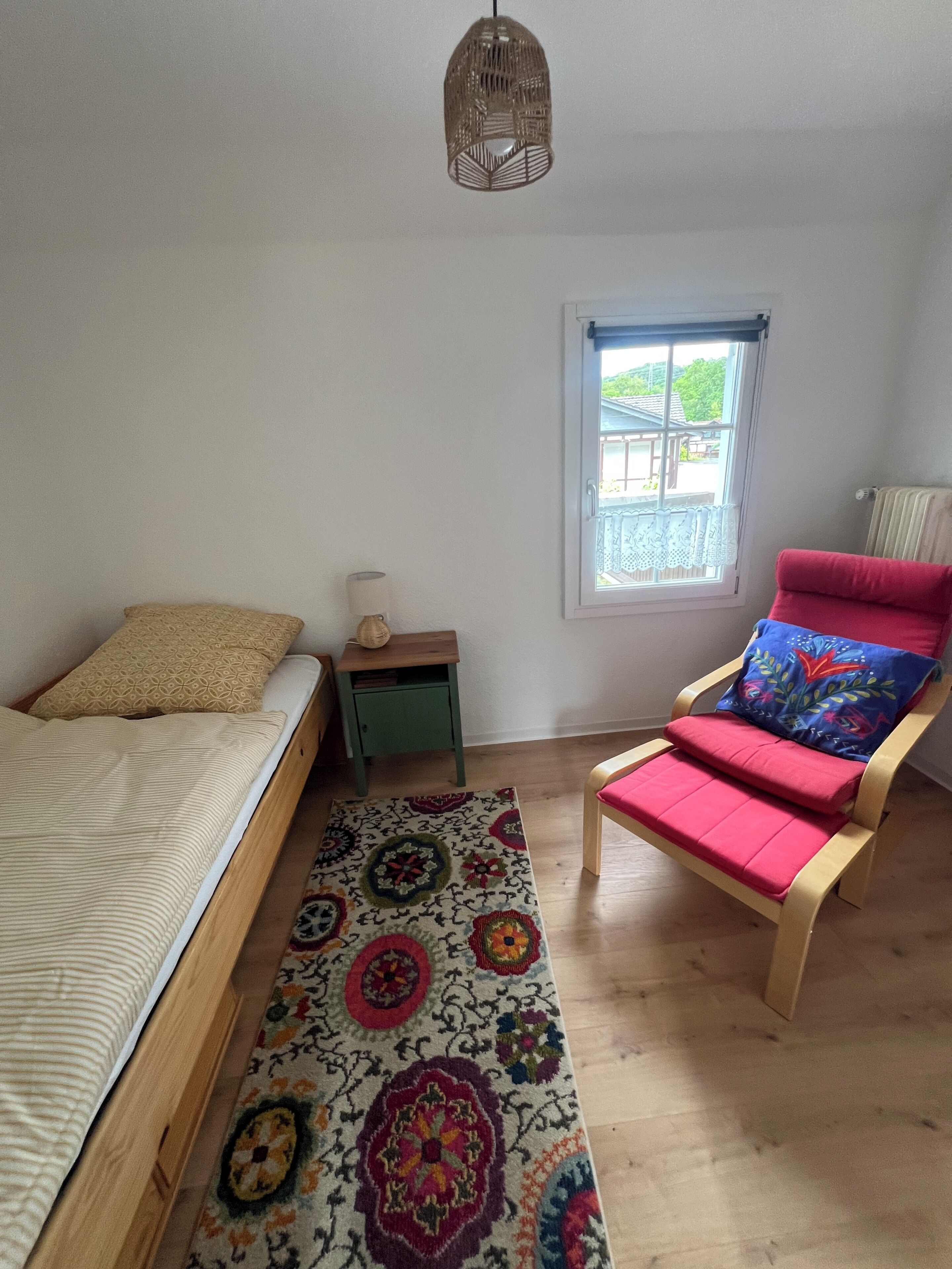 Une chambre à coucher comprenant un lit, une chaise, une petite table, une fenêtre et un luminaire suspendu.