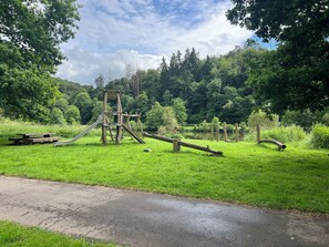 A playground with a slide and climbing structure, surrounded by lush greenery and a body of water.