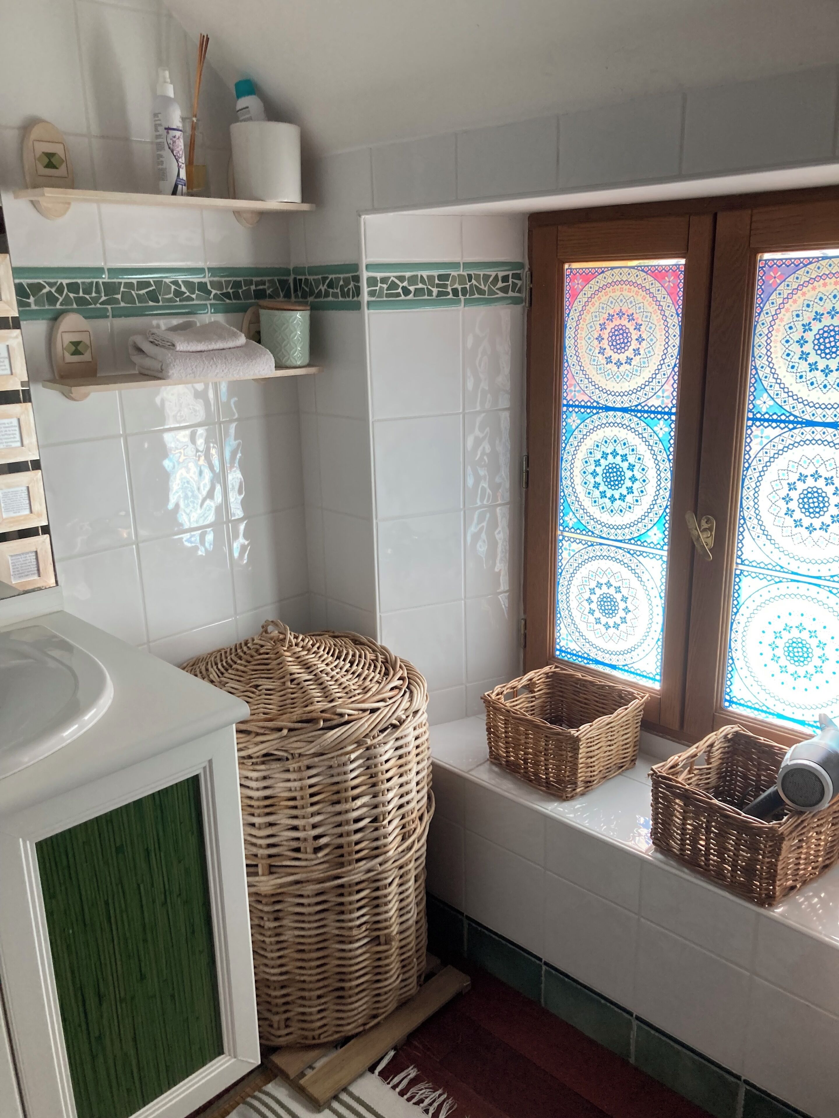 Une salle de bain aux carreaux blancs, avec un mur d’accent vert, des paniers en osier et une fenêtre ornée d’un motif coloré.