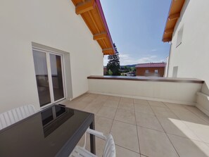 Ein Balkon mit Glastisch und Stühlen, Blick auf ein rotdachiges Gebäude und einen klaren Himmel.