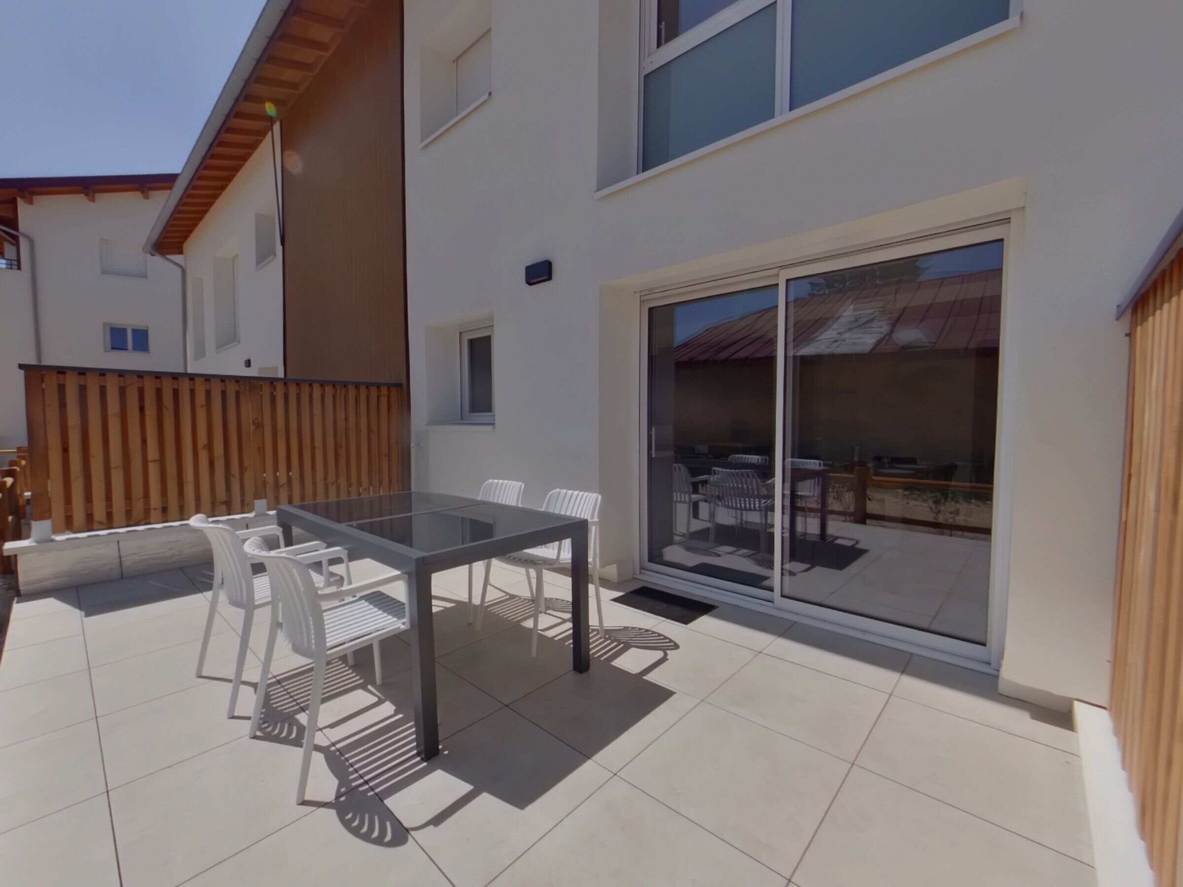 Eine moderne Terrassenanlage im Freien mit einem Glastisch und Stühlen, einem Holzzaun und einer Schiebetür zu einem angrenzenden Raum.