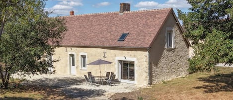 A stone house with a tiled roof, a patio with chairs and an umbrella, and a small garden area.