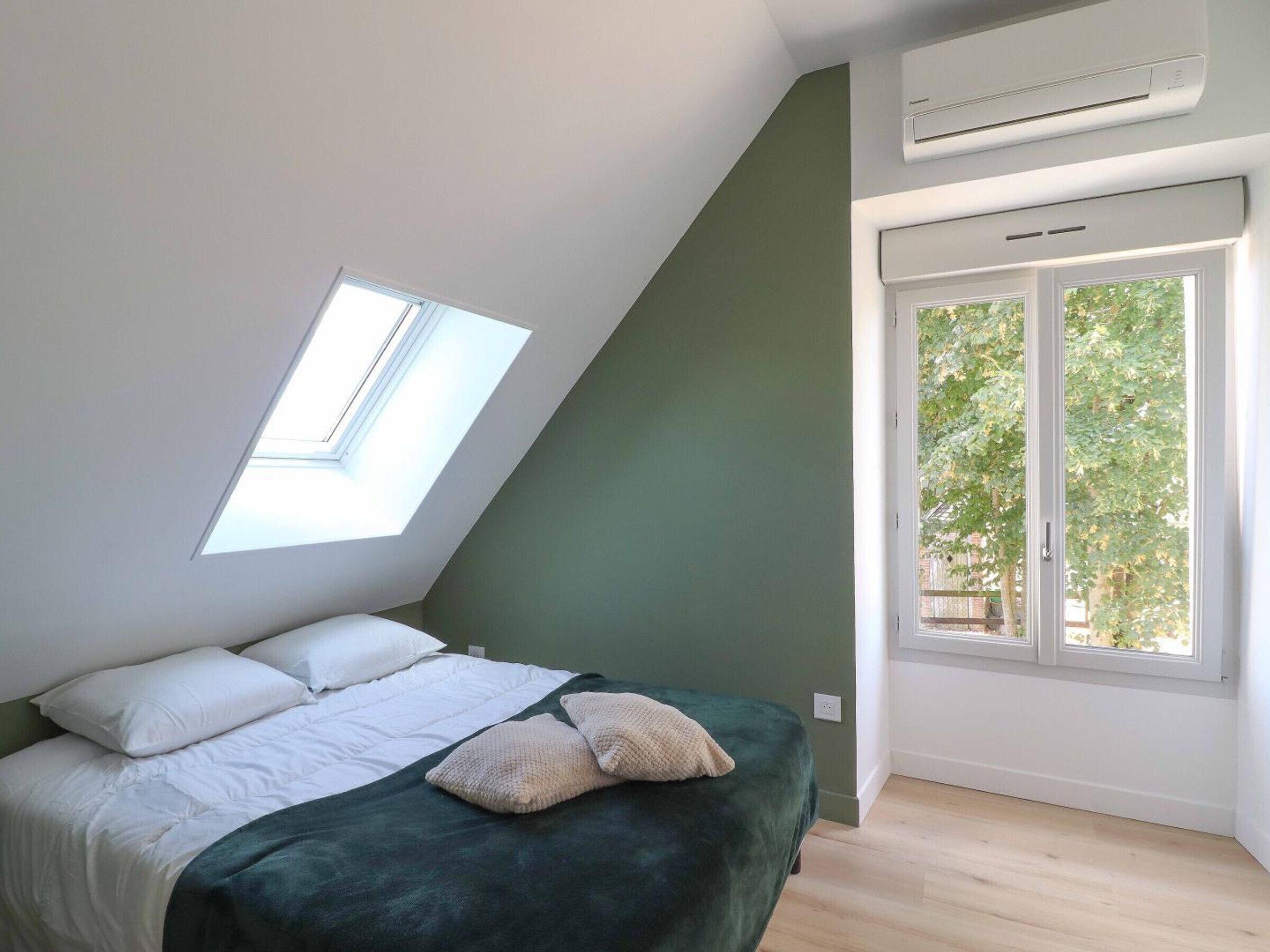 Een slaapkamer met dakraam, een bed met wit beddengoed, een groene accentmuur en een raam met uitzicht op groen.